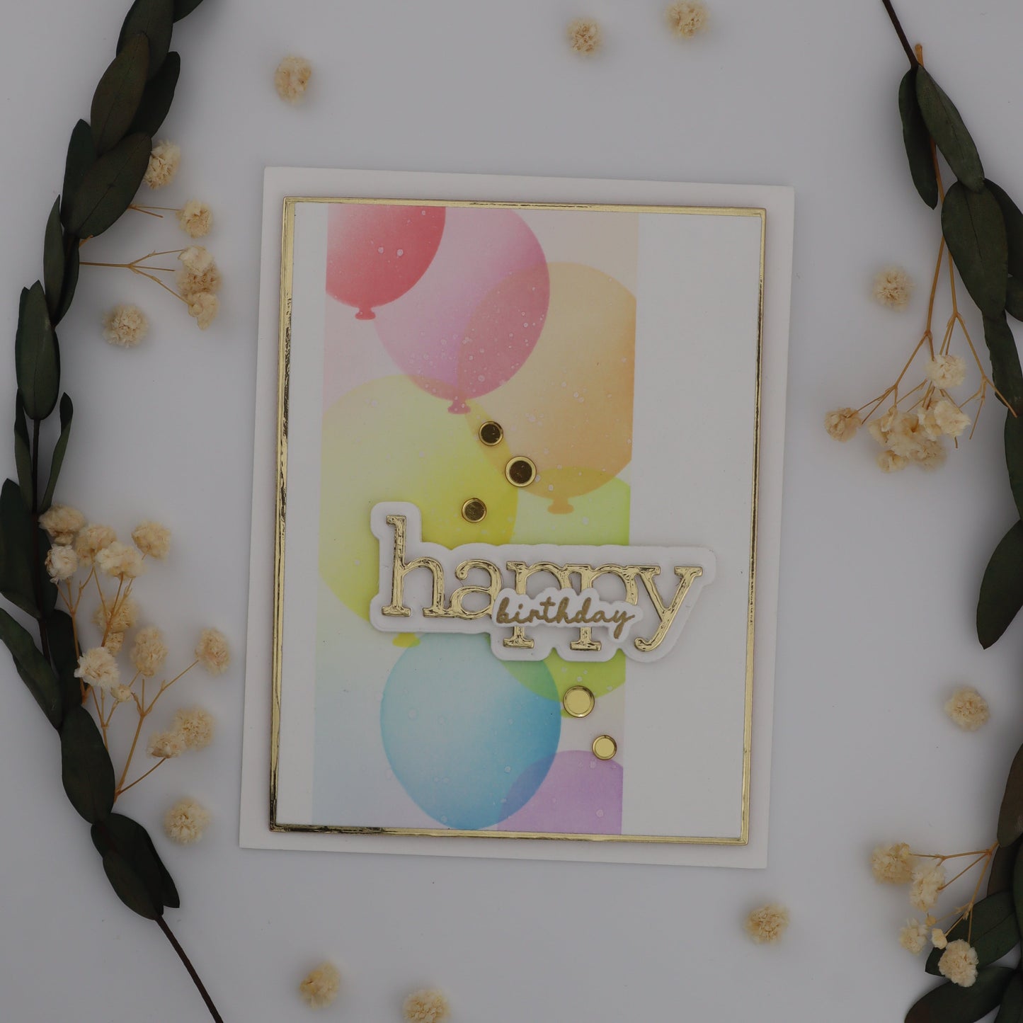 Decorative card with colorful balloons and 'Happy' text, surrounded by greenery and small flowers on a white background.