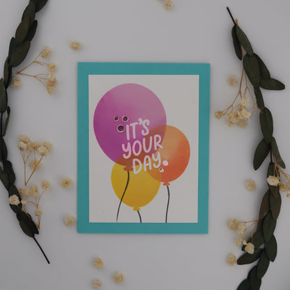 Colorful card with balloons and 'It's Your Day' text on a white background with decorative elements.