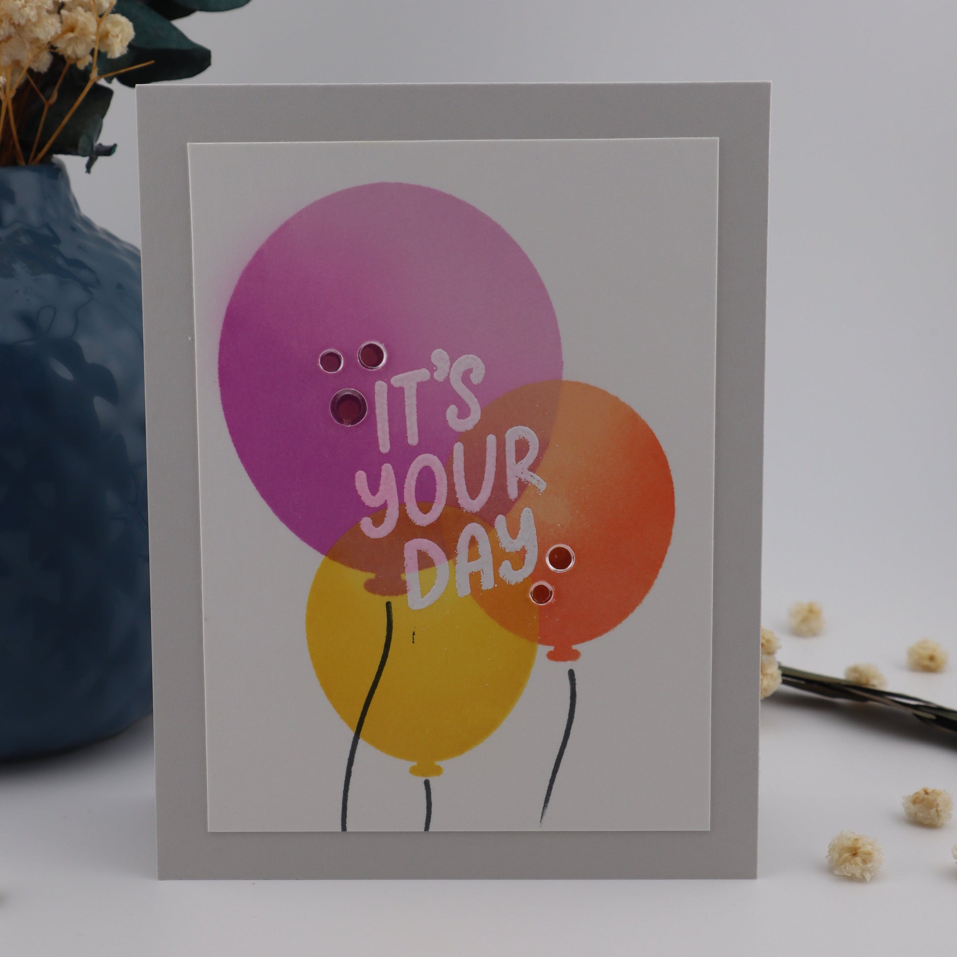 Card with balloons and 'It's Your Day' text on a light background with flowers.