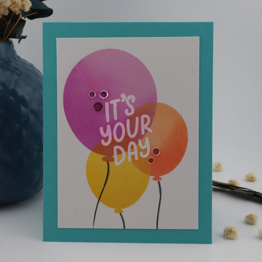 Greeting card with colorful balloons and 'It's Your Day' text on a white background with decorative elements.