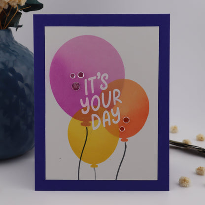 Greeting card with colorful balloons and 'It's your day' text on a white background.