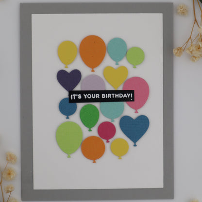 Framed card with colorful balloons and 'It's Your Birthday!' text on a white background with flowers.