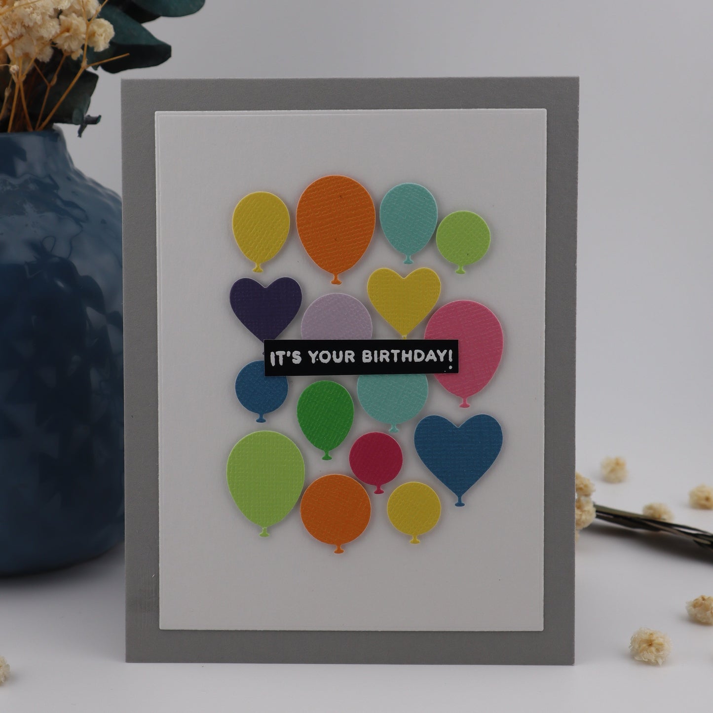 Framed card with colorful balloon design and 'It's Your Birthday' text on a white background with decorative elements.