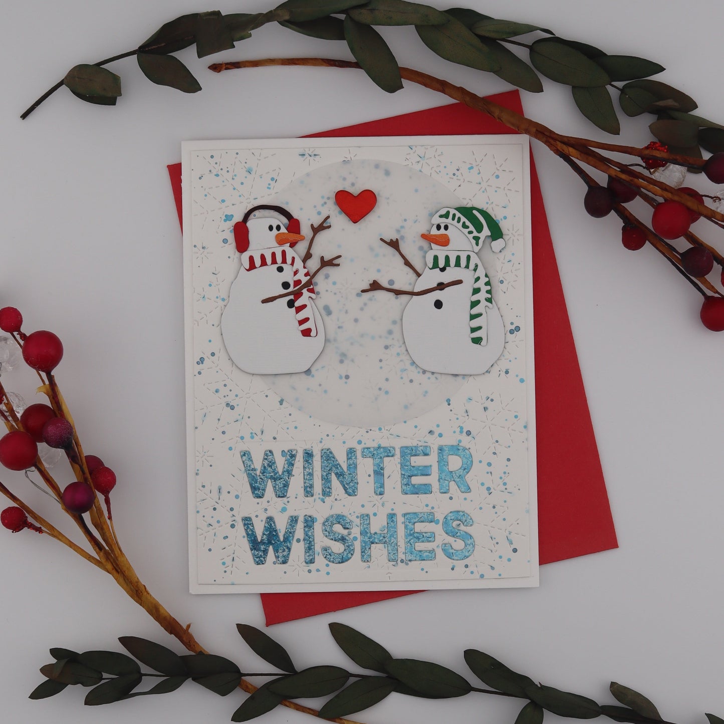 Card with snowmen and 'Winter Wishes' text, surrounded by greenery and red berries on a white surface.
