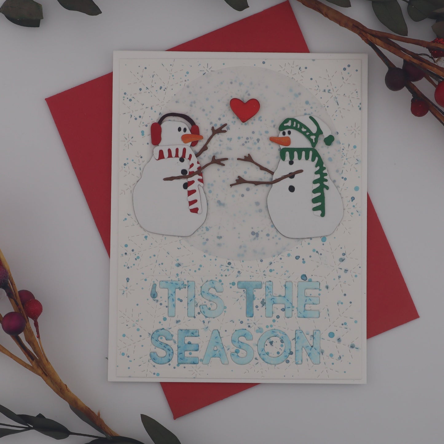 Christmas card with snowmen and 'Tis the Season text, surrounded by red berries and greenery on a white background.