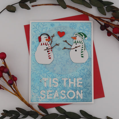 Christmas card with snowmen and 'Tis the Season text, surrounded by decorative branches on a white background.