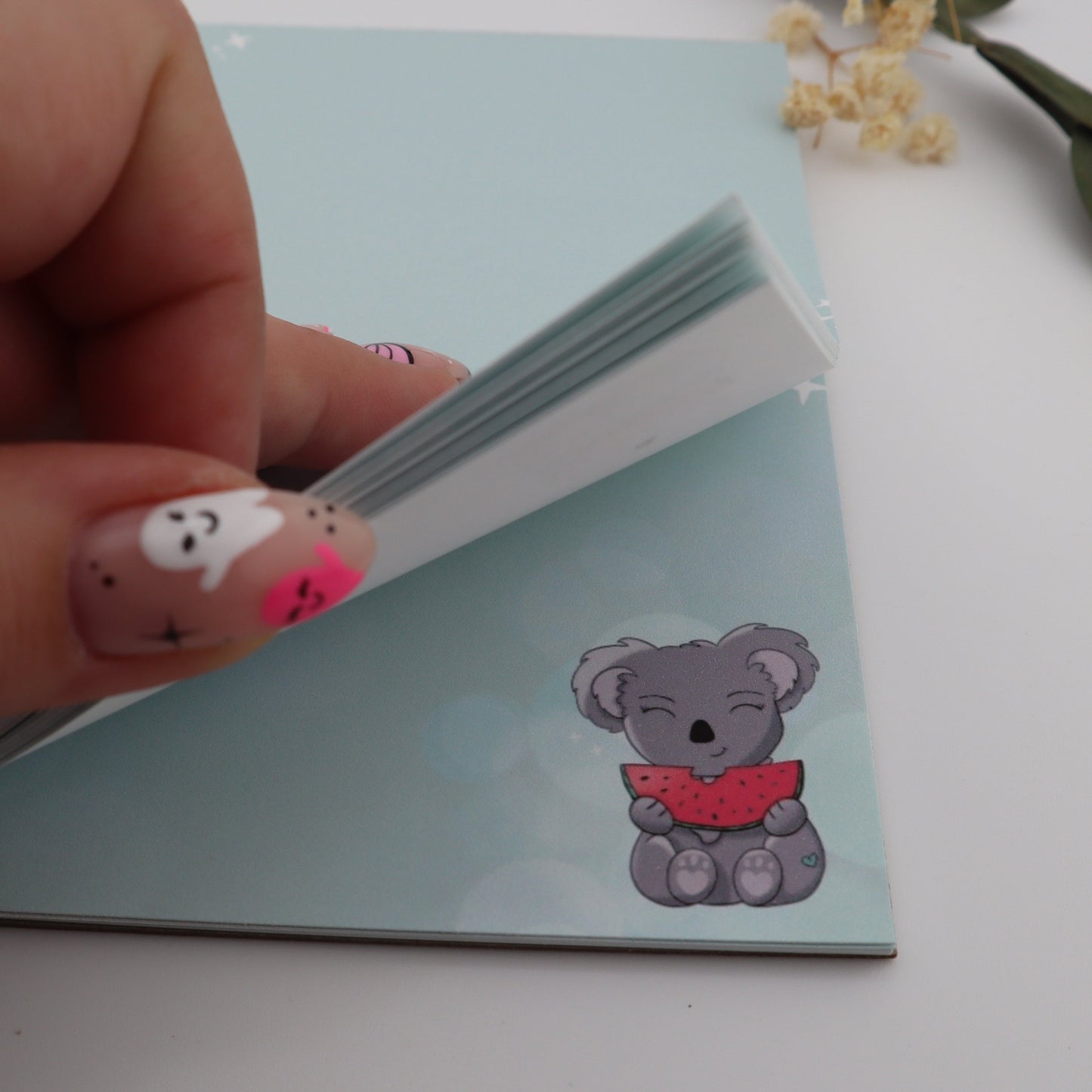 Hand holding a small card with a bear design on a light surface with dried flowers.