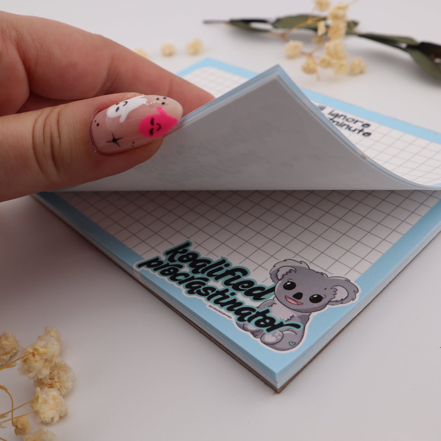 Hand holding a small notepad with a bear design on a white surface with dried flowers.