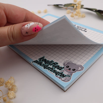 Hand holding a small notepad with a bear design on a white surface with dried flowers.